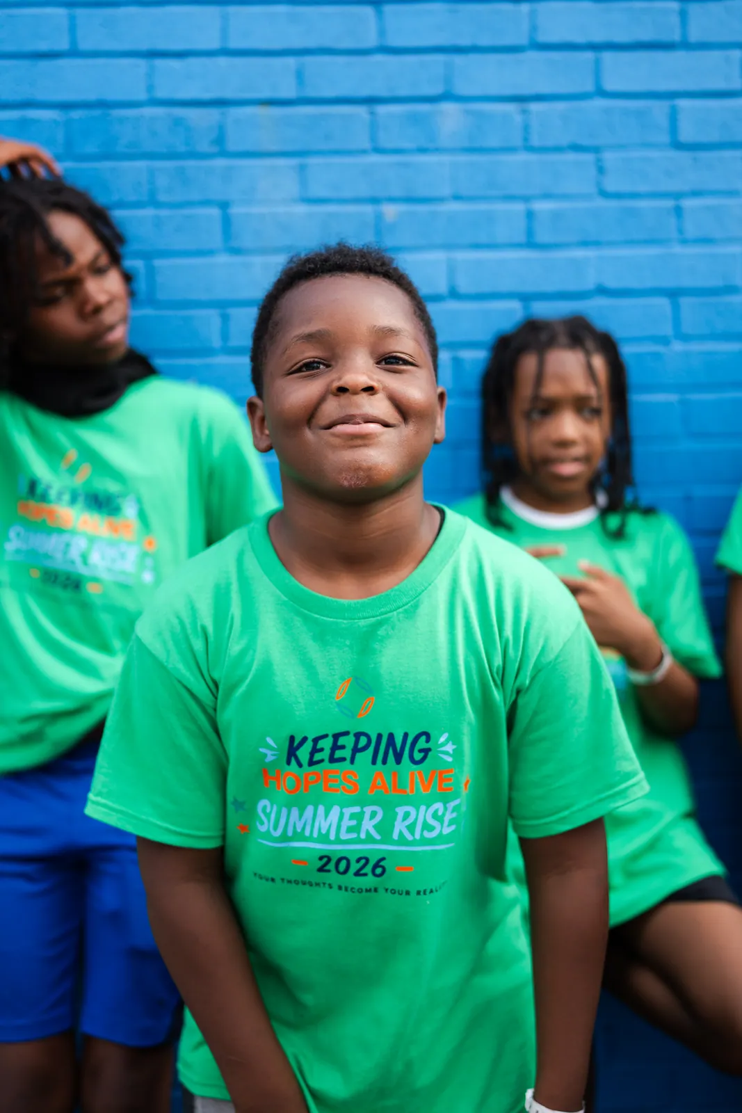 A smiling boy in a Summer RISE 2026 shirt against a blue wall